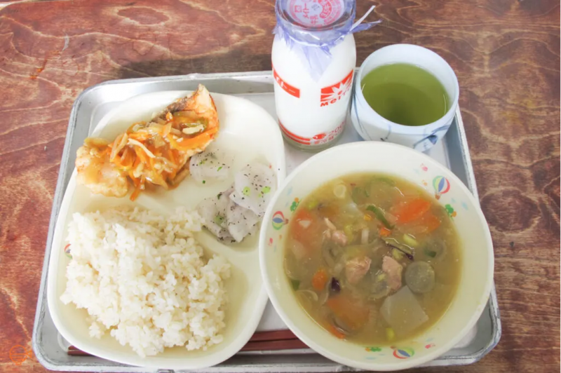 A serving of rice, battered fish with a sweet and sour topping, plus a side of pickles. To the right is a bowl of miso based pork and vegetable soup, and a bottle of milk and cup of green tea.