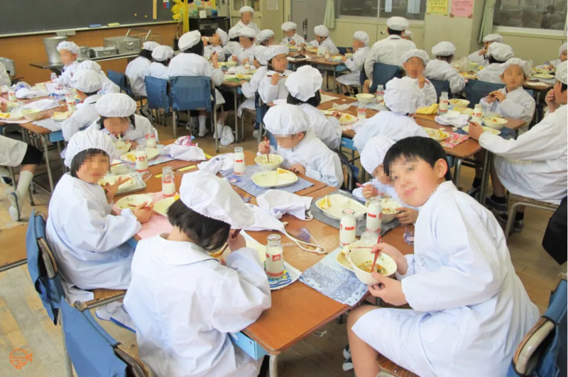 An elementary school class with their desks arranged into small groups, happily chat while eating school lunch, with a couple of them turning to pose for the camera.